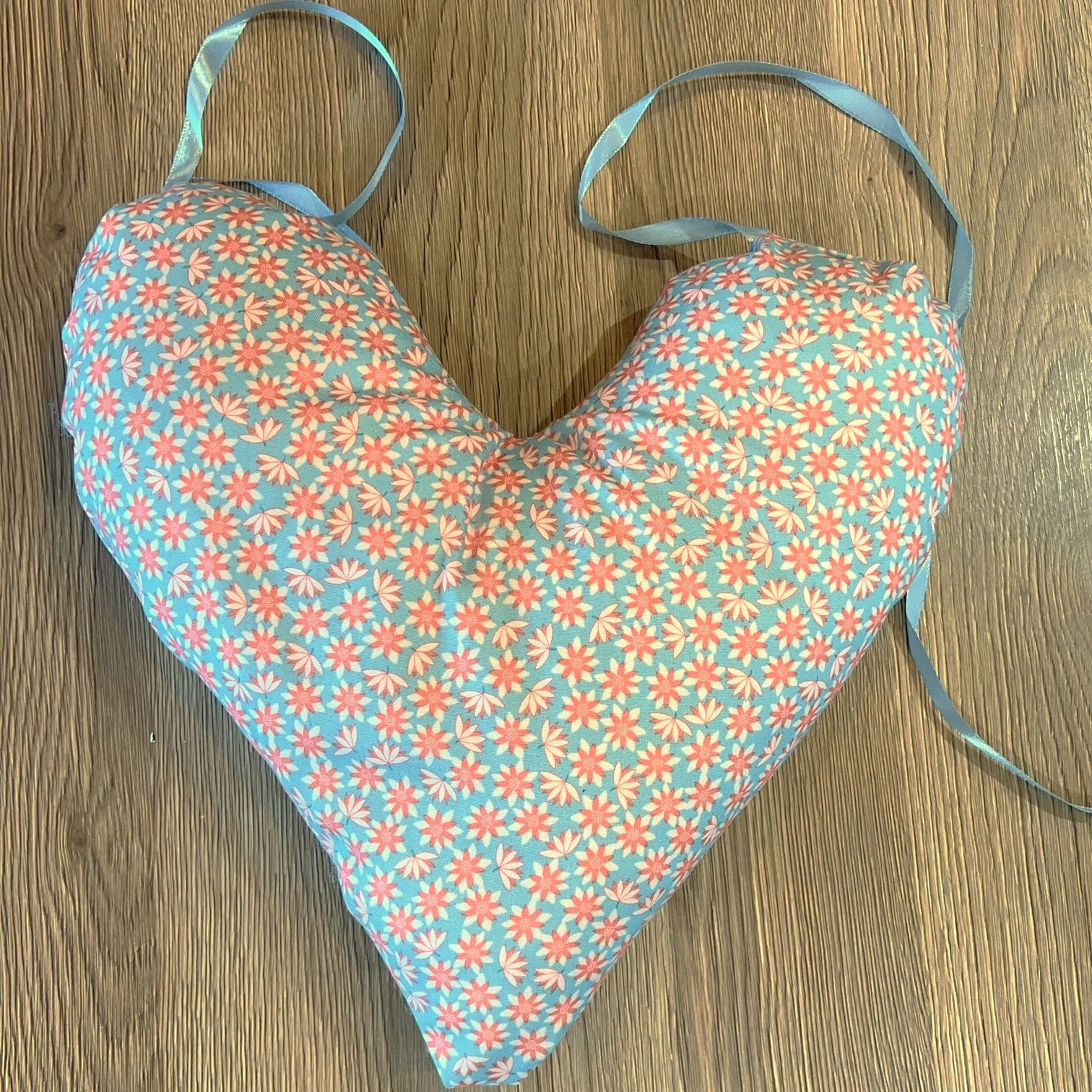 A handmade heart shaped mastectomy pillow with a light blue floral pattern and matching light blue straps, laid flat on a wooden floor.