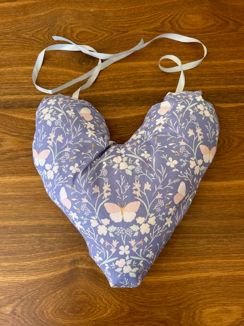 A handmade heart shaped mastectomy pillow with a lavender floral and butterfly pattern, featuring a white tie-on ribbon, laid flat on a wooden surface.