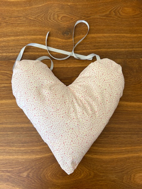 A handmade heart shaped mastectomy pillow made of light pink fabric with a tiny white floral print, featuring a white tie-ribbon strap, laid flat on a brown wooden surface.