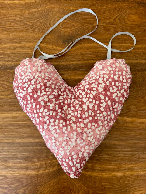 A handmade heart shaped mastectomy pillow with a pink and white speckled pattern, featuring white elastic straps, laid flat on a brown wooden surface.
